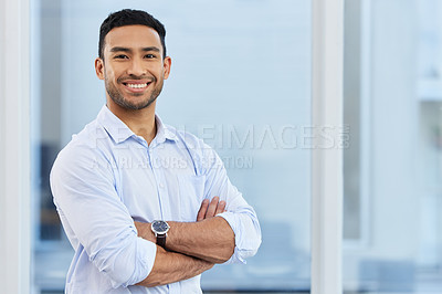Buy stock photo Shot of a handsome young businessman standing alone in his office with his arms folded
