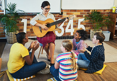 Buy stock photo Full length shot of a young teacher sitting and playing the guitar for her students in her classroom at school