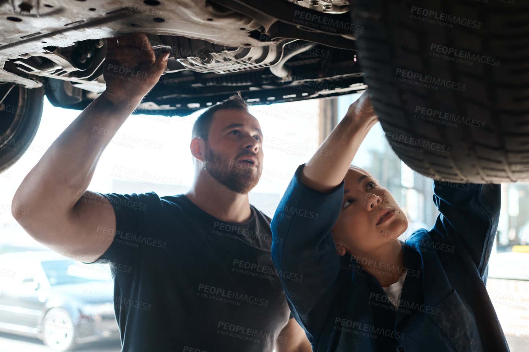 Buy stock photo Mechanic, garage and working on car wheel with training, maintenance engineer and teamwork at workplace. Repair shop, trade and mentor with woman checking under motor with technician and auto service