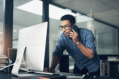 Buy stock photo Shot of a young man using his cellphone while working on a computer in a modern office