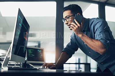 Buy stock photo Shot of a young man using his cellphone while working on a computer in a modern office