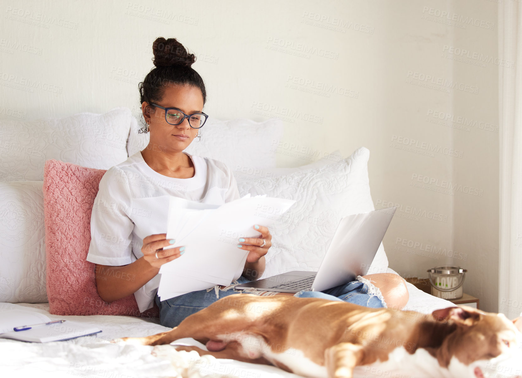 Buy stock photo Shot of a beautiful young woman using a laptop and completing paperwork with her dog in bed at home