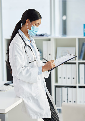 Buy stock photo Shot of a doctor wearing a face mask and standing alone while writing on her clipboard in her clinic