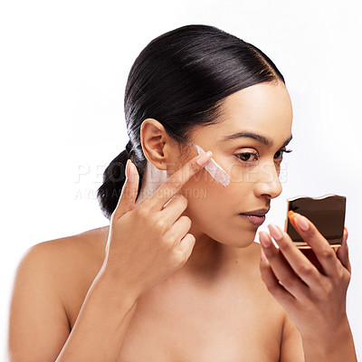 Buy stock photo Studio shot of a beautiful young woman using a mirror while applying moisturiser to her face against a white background