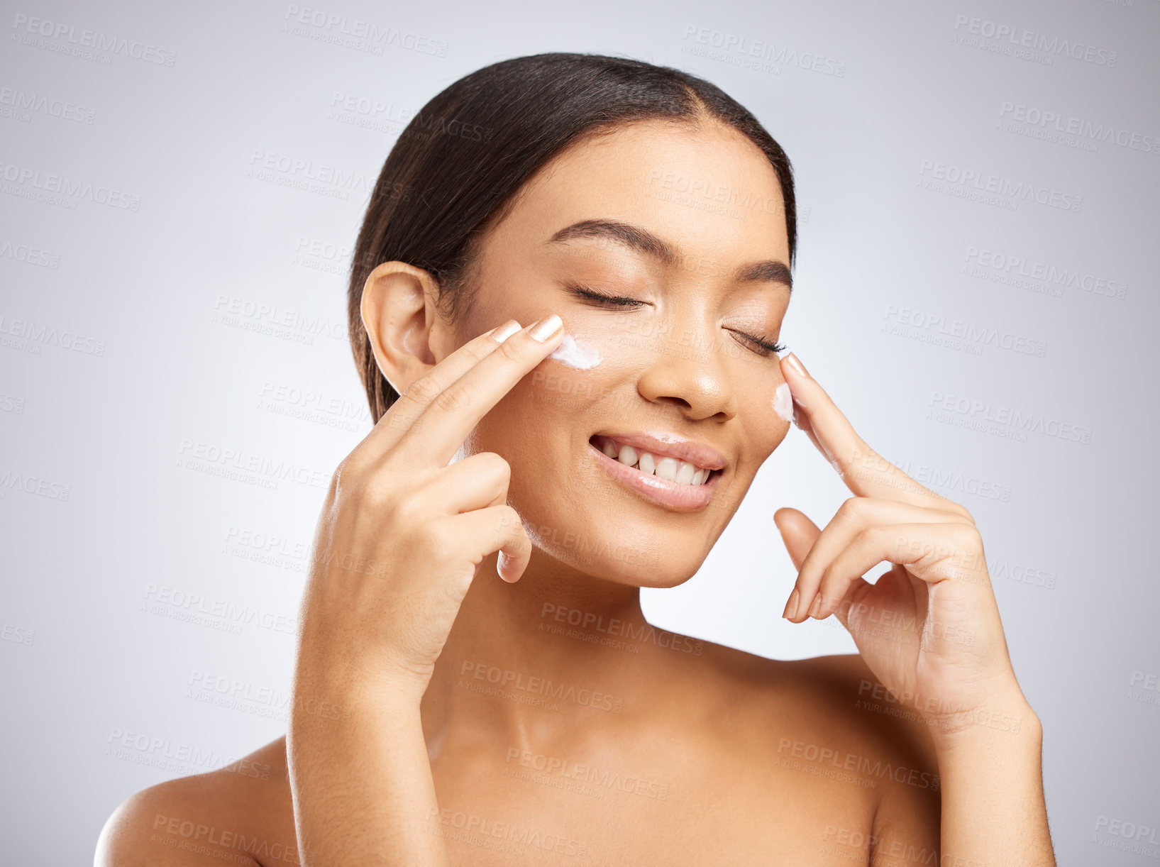 Buy stock photo Studio shot of an attractive young woman applying moisturiser to her face against a grey background