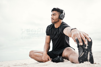 Buy stock photo Shot of a young man wearing headphones while stretching on the beach