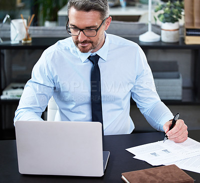 Buy stock photo Shot of a mature businessman using a laptop while going through paperwork in an office at work