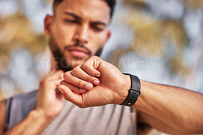 Buy stock photo Shot of a young man standing alone outside and using his watch to time his pulse after a run