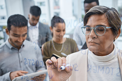 Buy stock photo Shot of a mature businesswoman writing notes on a glass screen while brainstorming with her colleagues in an office