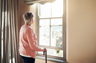 Buy stock photo Shot of a senior woman with a cane looking thoughtfully out of a window at home