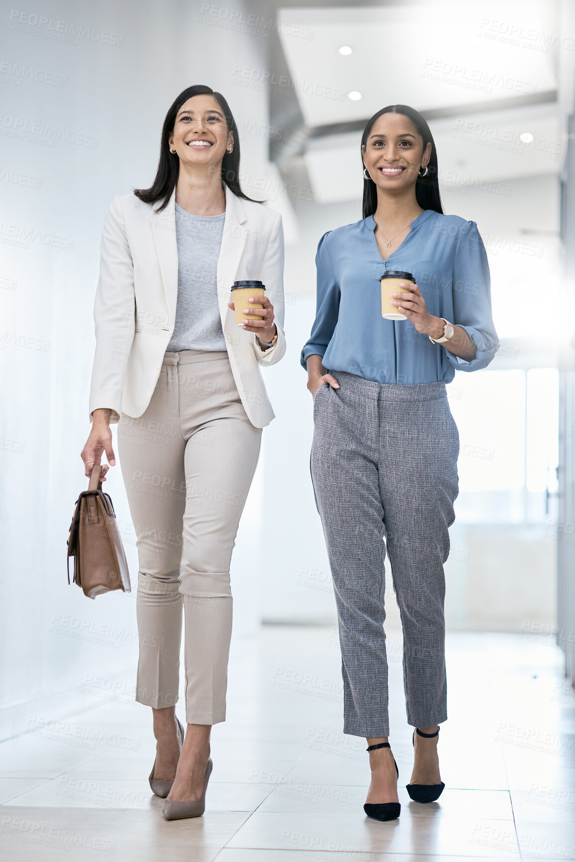 Buy stock photo Shot of two businesswomen carrying coffees while walking through an office