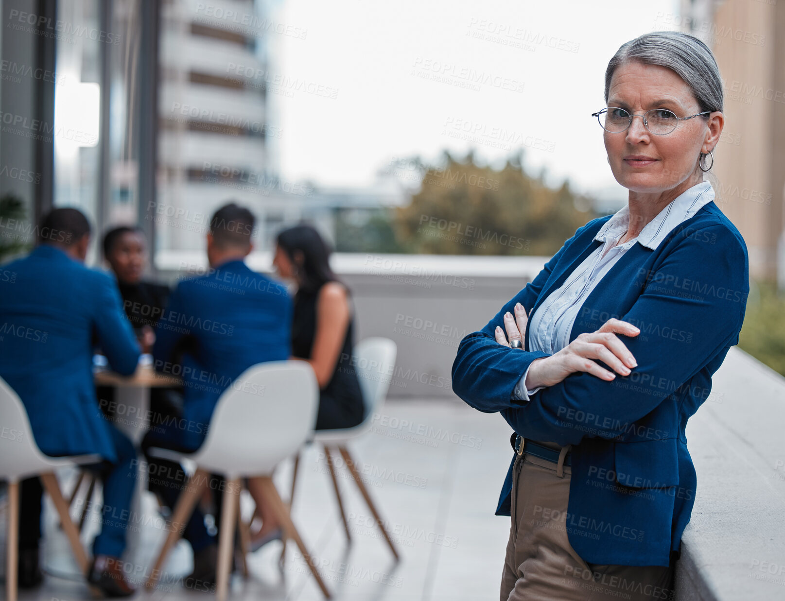 Buy stock photo Cropped portrait of an attractive mature businesswoman standing with her arms folded outside on the office balcony with her colleagues in the background