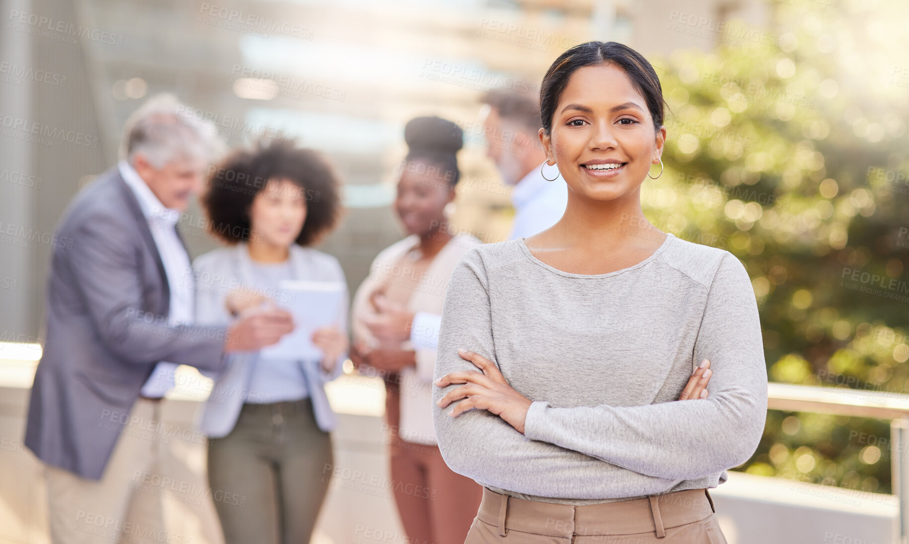 Buy stock photo Shot of an attractive young businesswoman standing outside with her arms folded while her colleagues discuss behind her