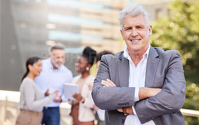 Buy stock photo Shot of a mature businessman standing outside with his arms folded while his colleagues discuss behind him