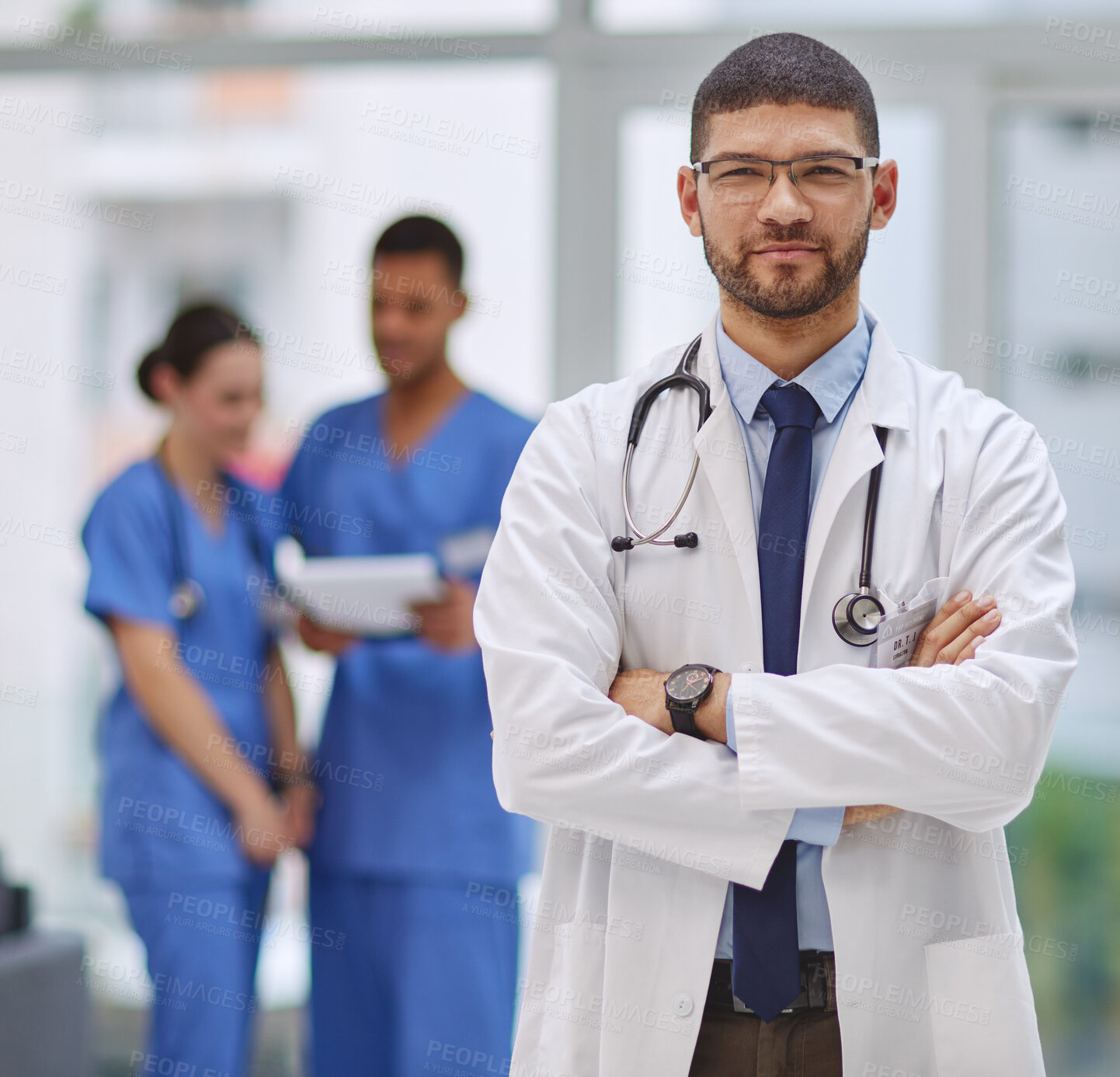 Buy stock photo Portrait of a young doctor standing with his arms crossed in a hospital with his colleagues in the background