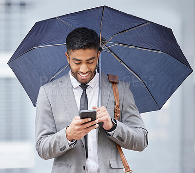 Buy stock photo Shot of a young businessman holding an umbrella while using a cellphone on a rainy day in the city