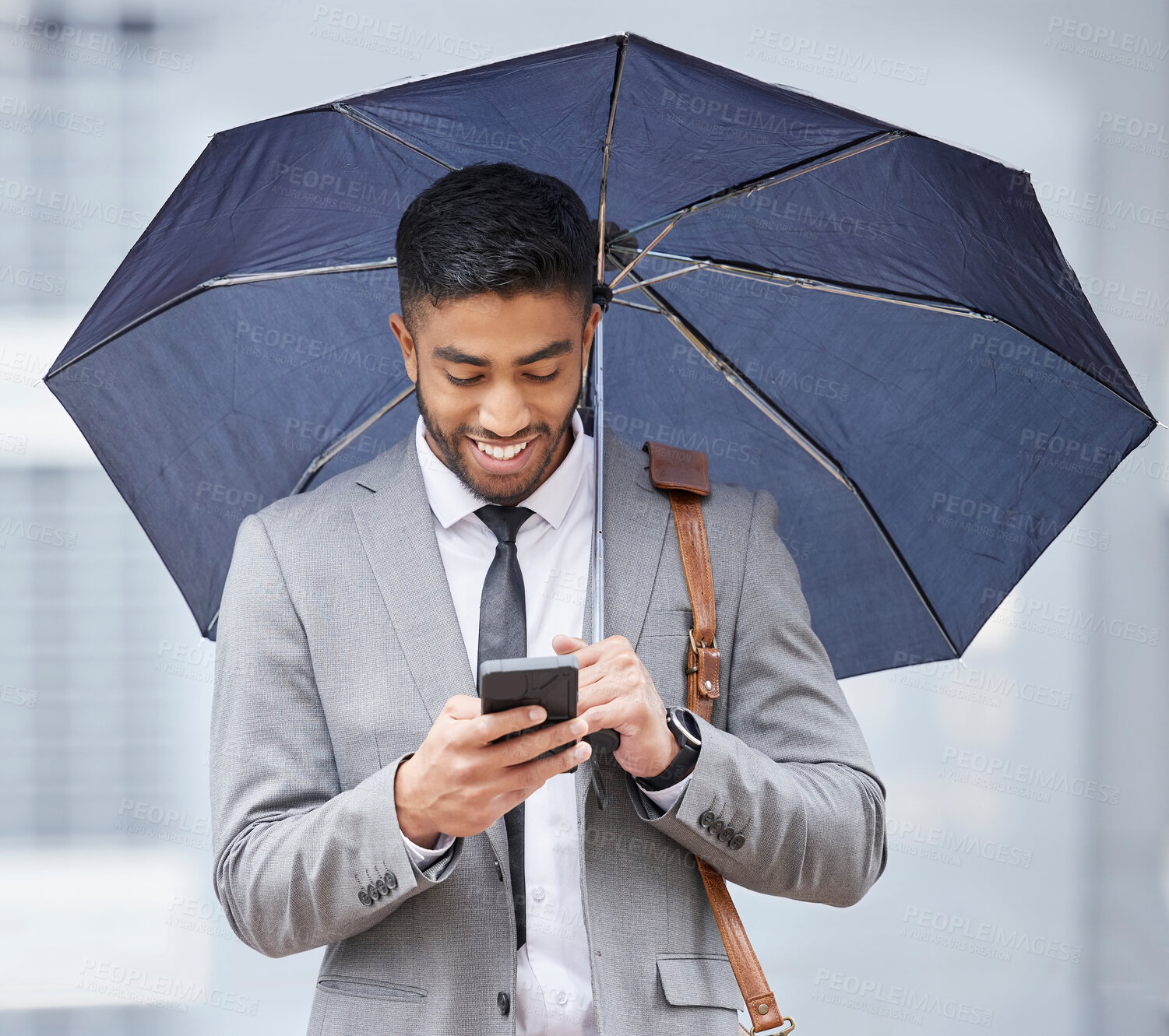 Buy stock photo Shot of a young businessman holding an umbrella while using a cellphone on a rainy day in the city