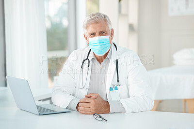 Buy stock photo Shot of a senior doctor sitting alone in his clinic and wearing a face mask while using his laptop