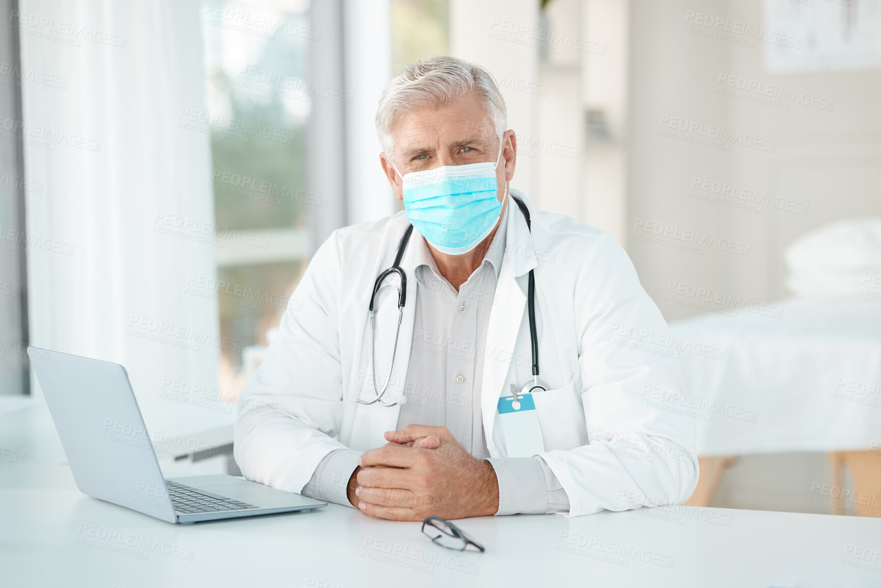 Buy stock photo Shot of a senior doctor sitting alone in his clinic and wearing a face mask while using his laptop