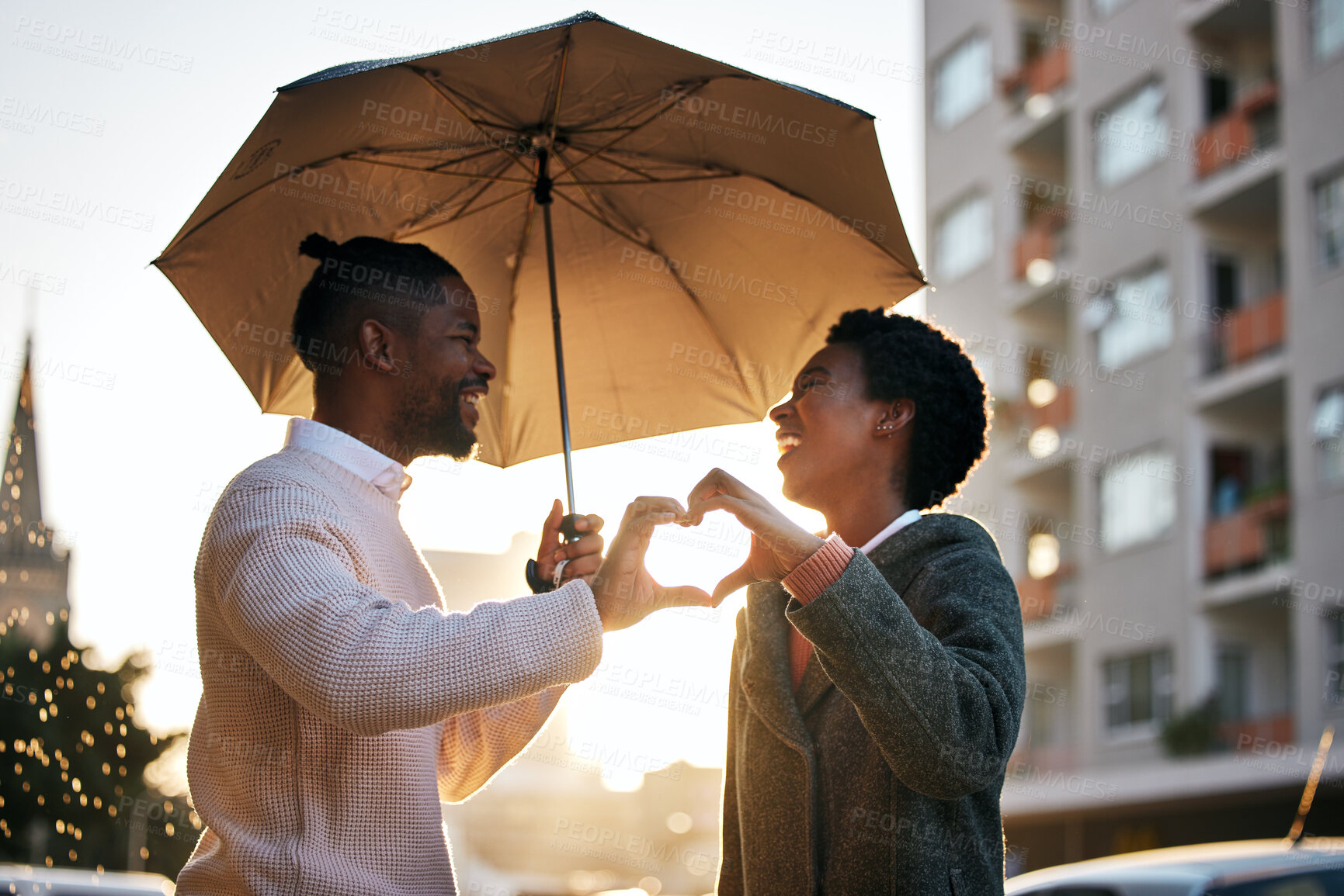 Buy stock photo Shot of a young man and woman standing under an umbrella and making a heart shaped gesture while going for a walk in the rain against an urban background