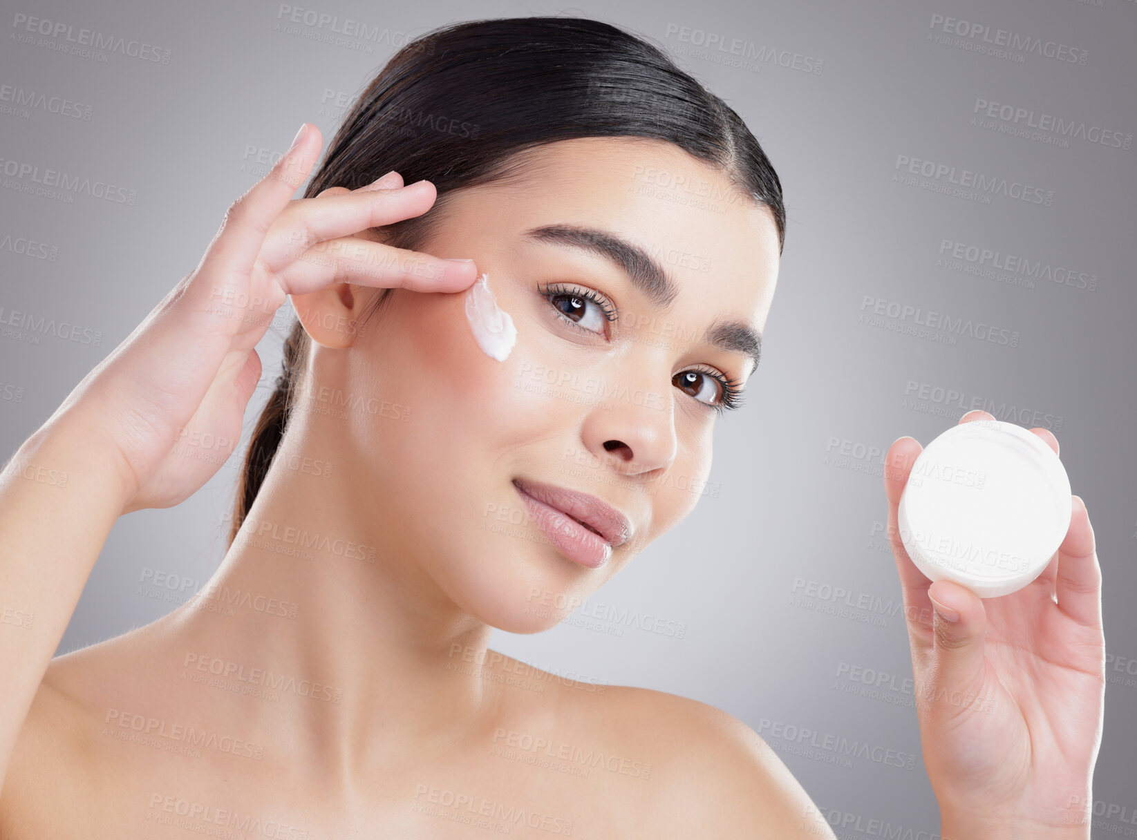 Buy stock photo Studio portrait of an attractive young woman applying lotion to her face against a grey background