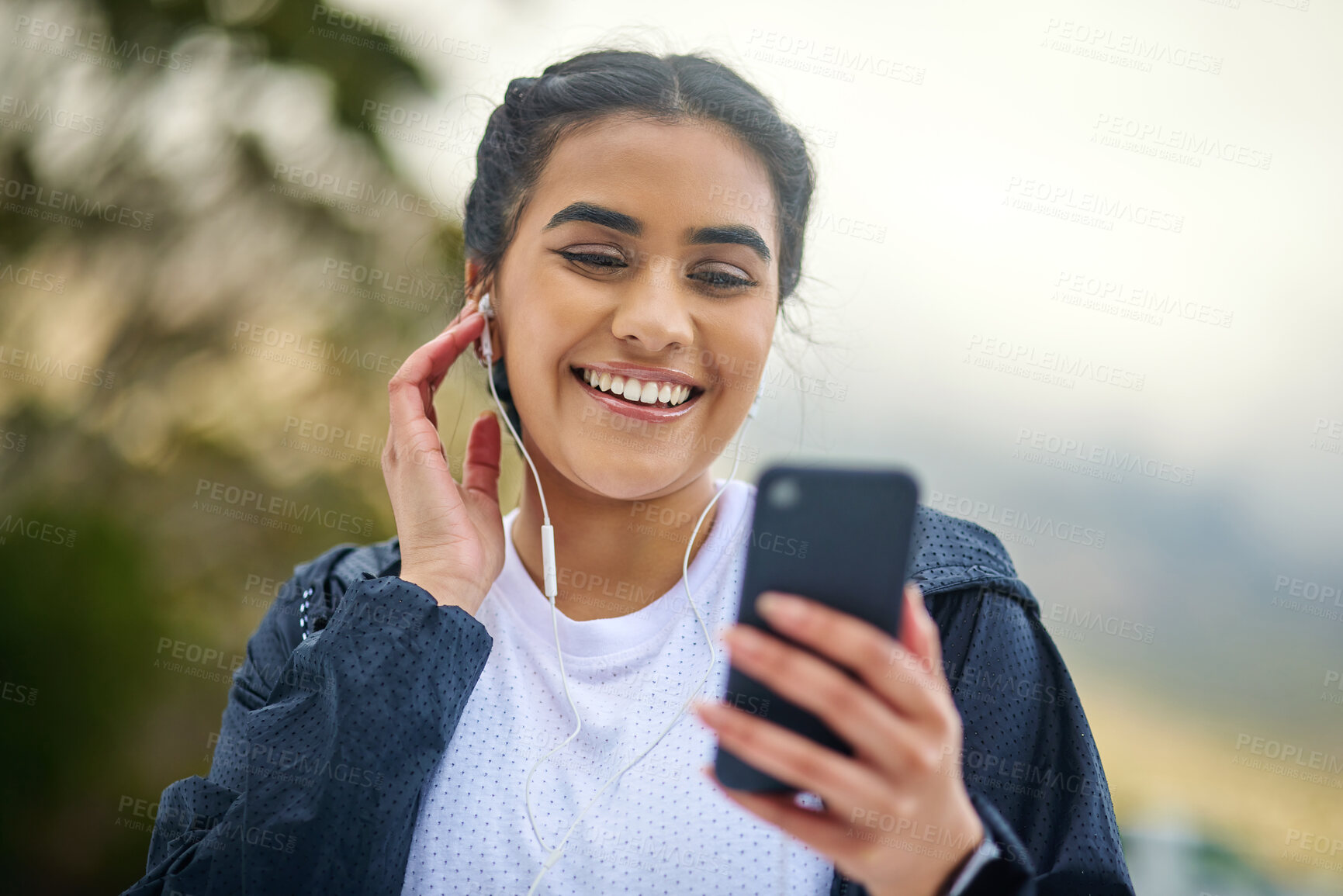 Buy stock photo Shot of a sporty young woman wearing earphones and using a cellphone while exercising outdoors