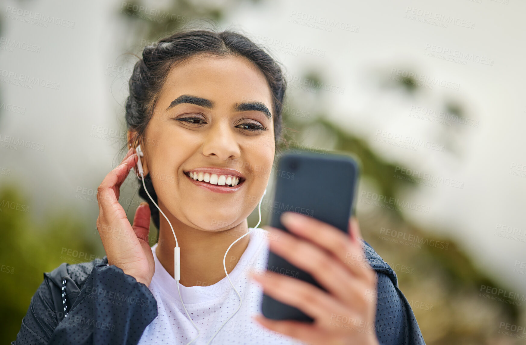 Buy stock photo Shot of a sporty young woman wearing earphones and using a cellphone while exercising outdoors