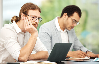 Buy stock photo Shot of a young businessman looking bored while working on a laptop in an office with his colleague in the background