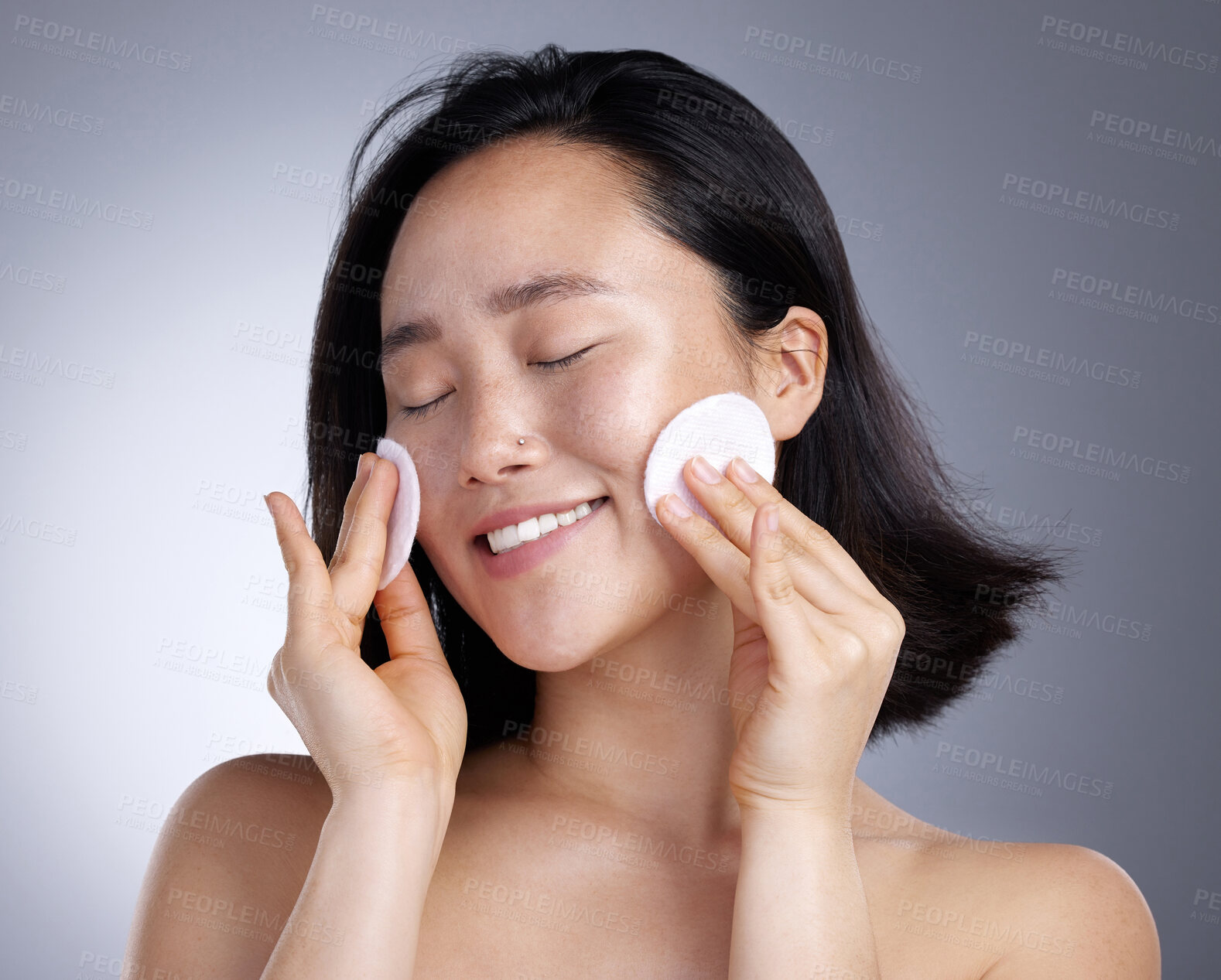 Buy stock photo Shot of a young woman using a cotton wipe on her face against a grey background