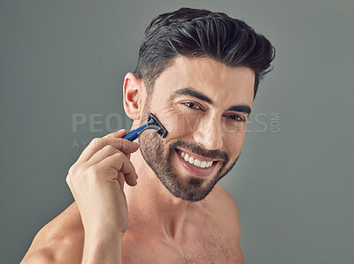 Buy stock photo Shot of a handsome young man shaving his beard while standing against a grey background