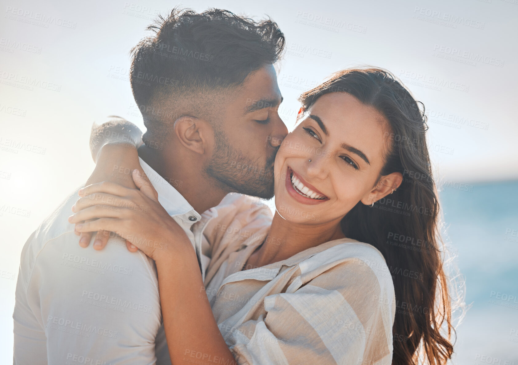 Buy stock photo Interracial, kiss or couple hug at beach for love on vacation, valentines day or holiday in Spain. Happy, relationship and people at sea for bonding together for romantic trip in ocean with smile