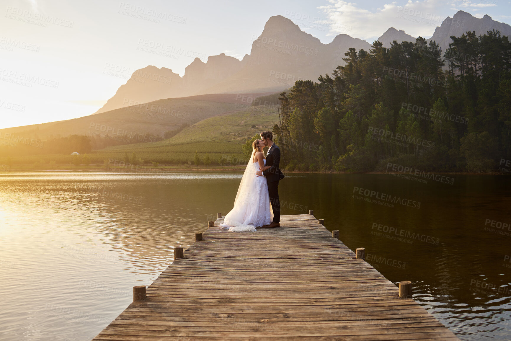 Buy stock photo Kiss, romantic and married couple by a lake for a wedding, marriage ceremony and event. Together, happy and a bride and groom standing by a pier, kissing and giving affection to celebrate their love