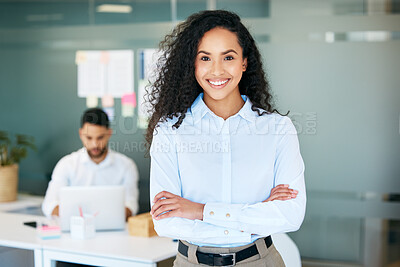 Buy stock photo Shot of an attractive young businesswoman standing in the office with her arms folded while her colleague works behind her