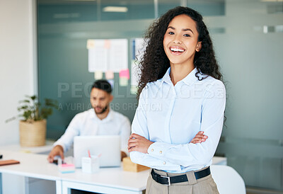 Buy stock photo Shot of an attractive young businesswoman standing in the office with her arms folded while her colleague works behind her