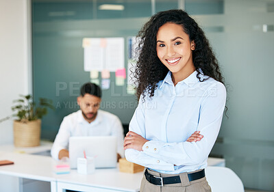 Buy stock photo Shot of an attractive young businesswoman standing in the office with her arms folded while her colleague works behind her