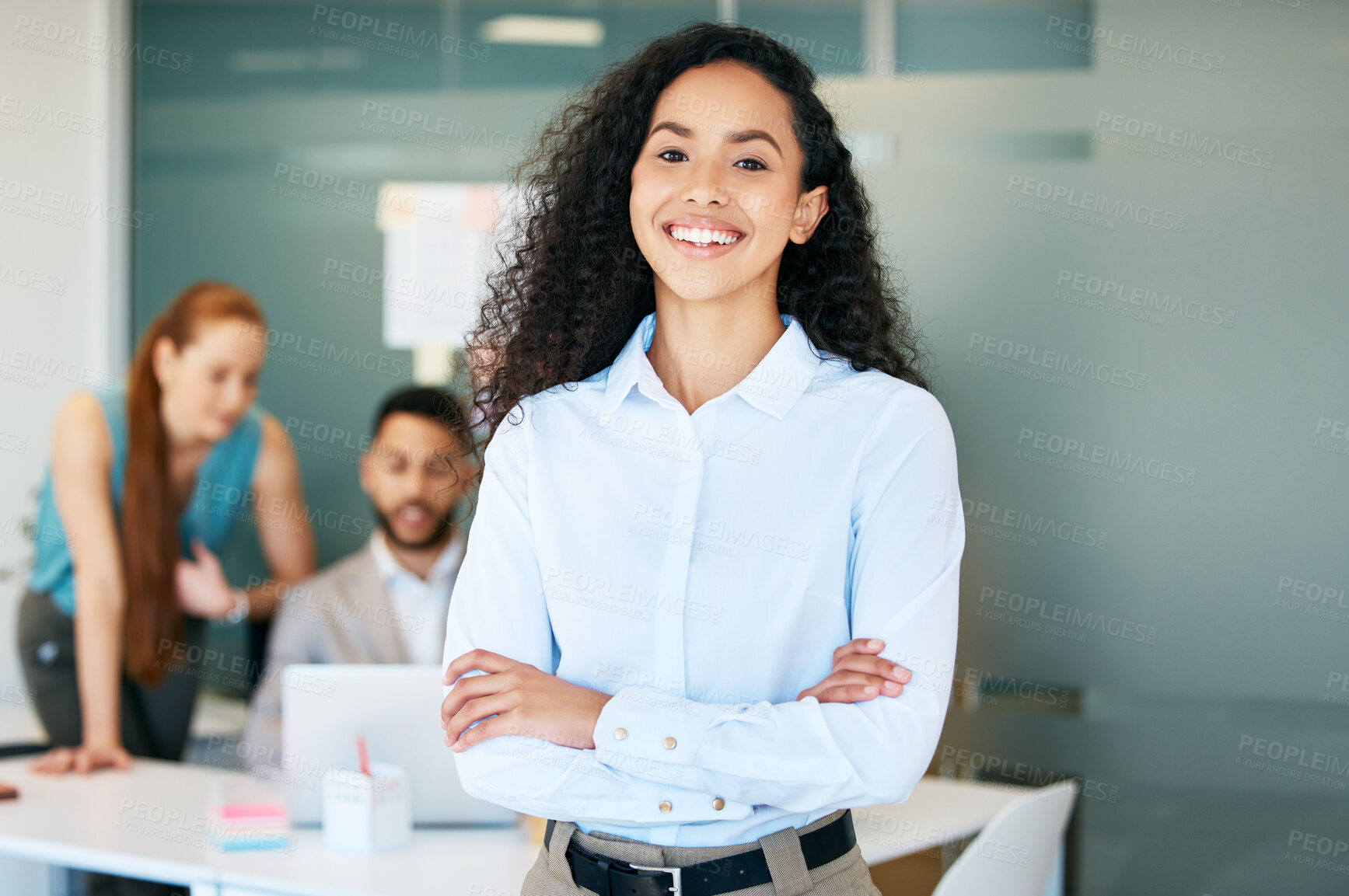 Buy stock photo Shot of an attractive young businesswoman standing in the office with her arms folded while her colleagues work behind her