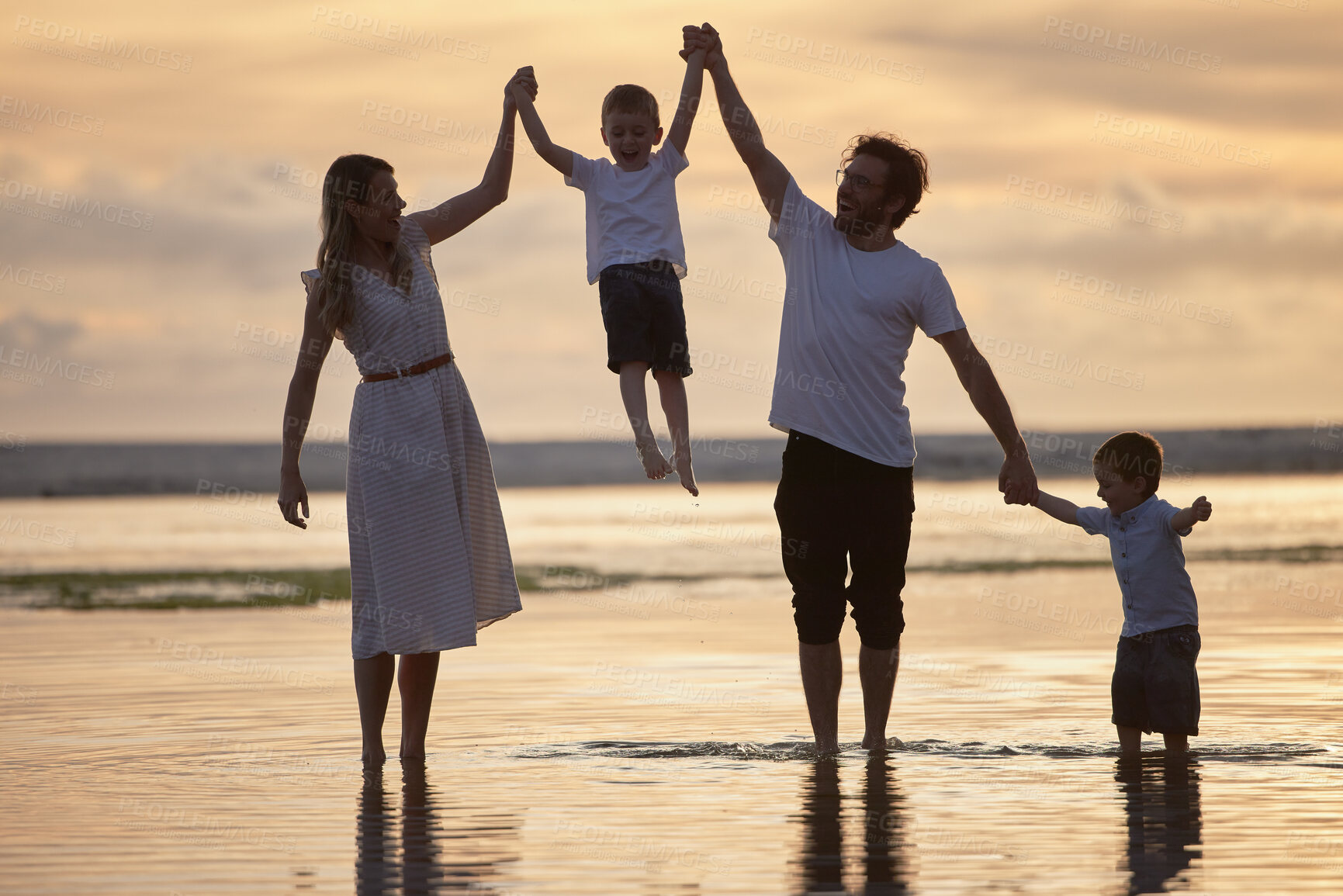 Buy stock photo Sunset, holding hands and family at beach on holiday, adventure or vacation together for bonding. Summer, fun and parents walking and lifting child in ocean with people on tropical weekend trip.