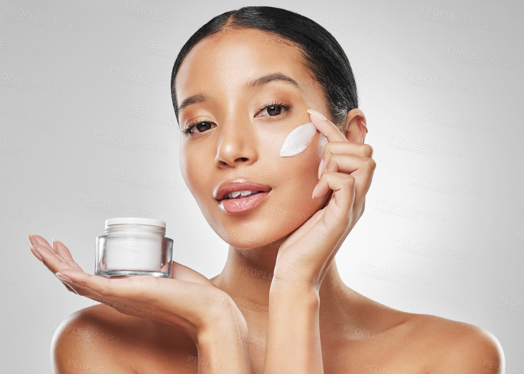 Buy stock photo Studio portrait of an attractive young woman applying moisturiser on her face against a grey background