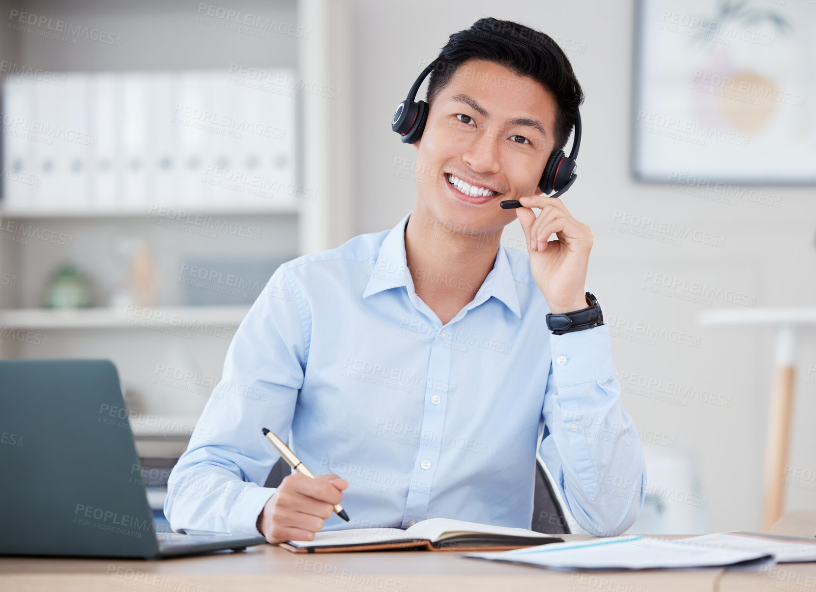 Buy stock photo Shot of a young male call center agent using a laptop and writing in a notebook at work