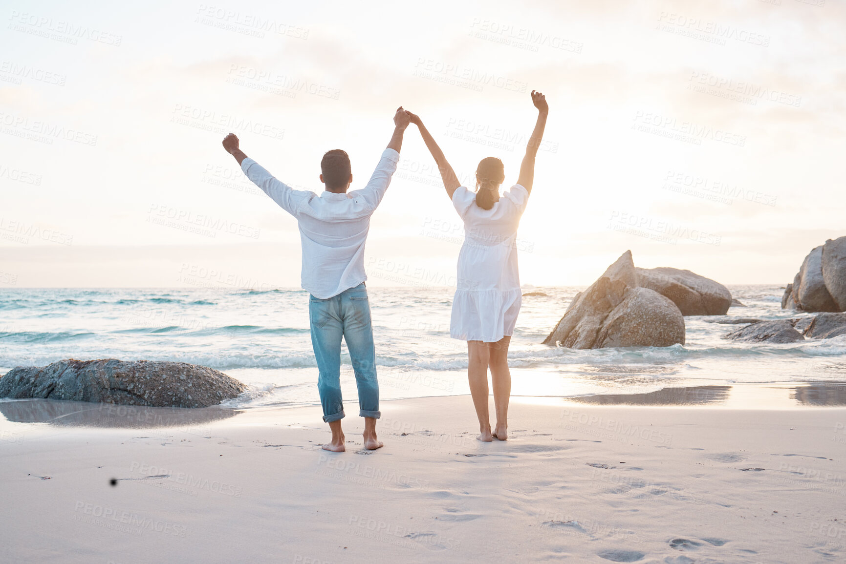 Buy stock photo Shot of a young couple standing with their hands raised at the beach