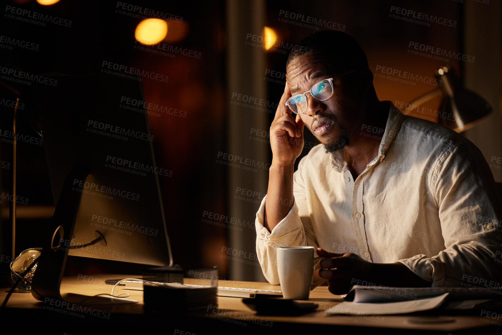 Buy stock photo Shot of a young businessman looking worried while drinking coffee and working on a computer in an office at night