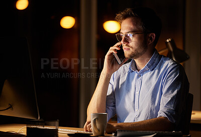 Buy stock photo Shot of a young businessman talking on a cellphone while working on a computer in an office at night