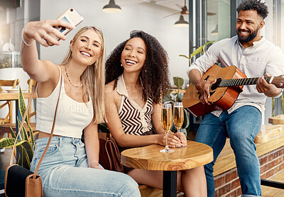 Buy stock photo Shot of two friends taking selfies together at a cafe while a man plays guitar