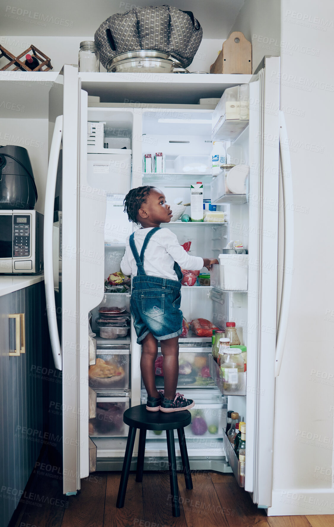 Buy stock photo Home, kid and food in fridge in kitchen with independence, hungry or checking for snack on chair. Black girl, child or search for meal, lunch or curious with growth and cognitive development in house