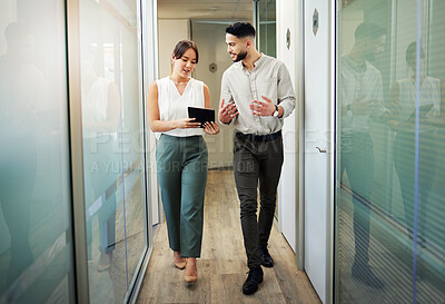 Buy stock photo Full length shot of two young businesspeople walking through the office and having a discussion while using a digital tablet
