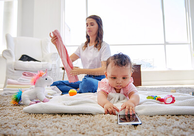 Buy stock photo Shot of a mother folding clothes while her baby plays in the lounge