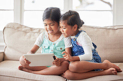 Two little girls playing online with a digital tablet, looking happy and content on the sofa at home