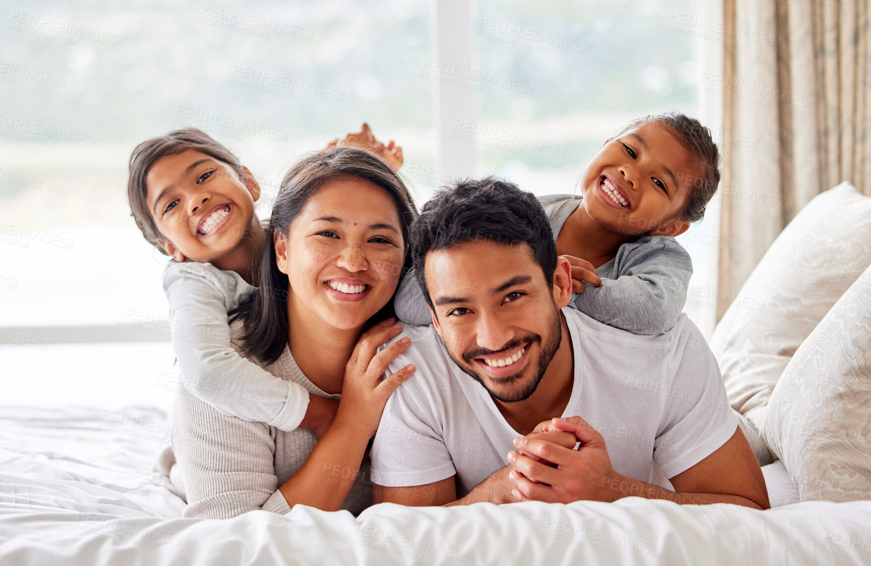 Buy stock photo Young happy smiling couple looking cheerful and relaxed while sharing the bed with their daughters. Two girls being playful while in bed with their parents