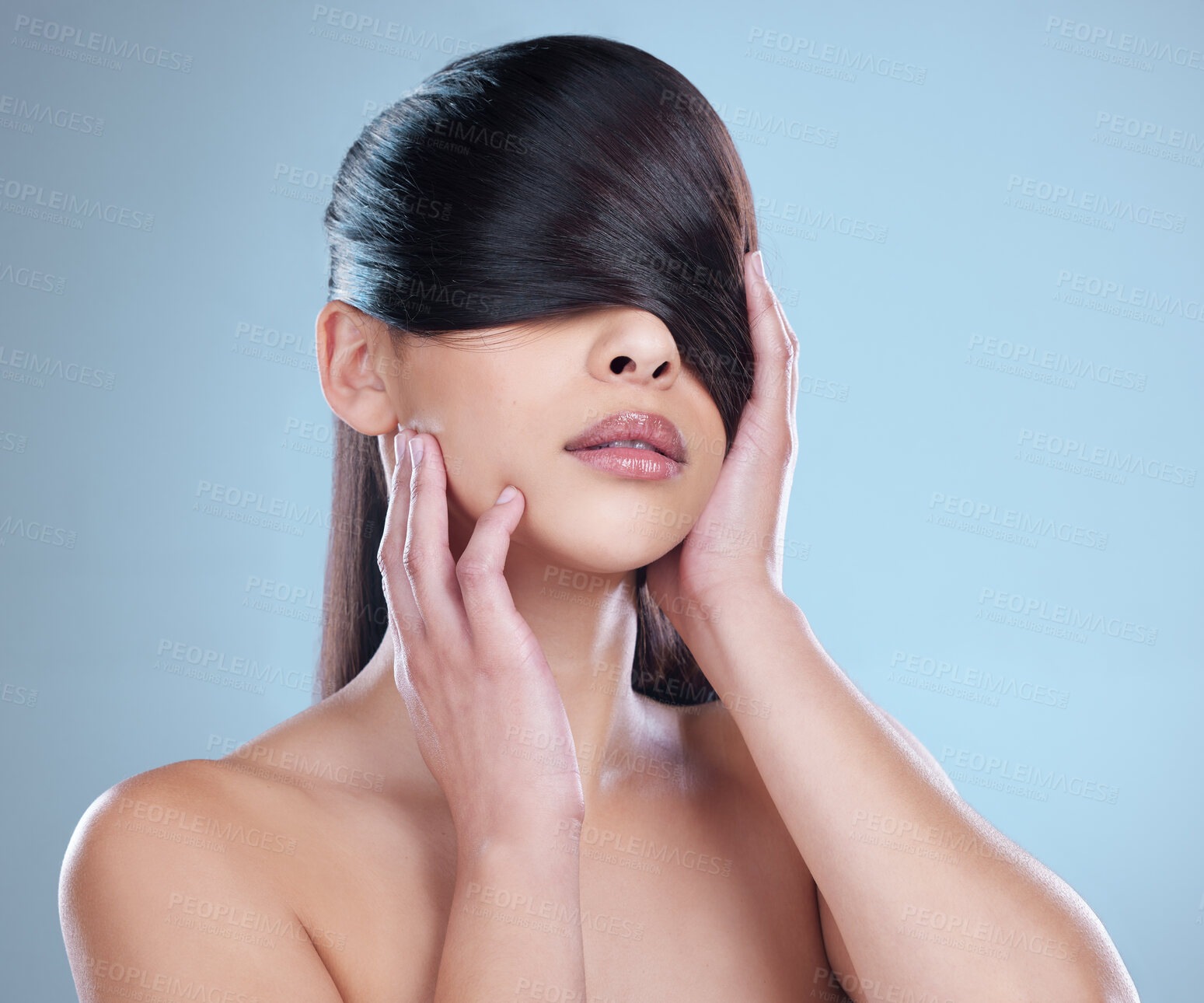 Buy stock photo Studio shot of a beautiful young woman playfully covering her eyes with her hair while posing against a blue background