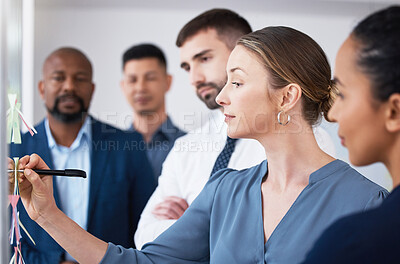 Buy stock photo Confident manager showing and teaching team of colleagues while writing on sticky notes on transparent board in office. Diverse group of business people brainstorming and planning a strategy together